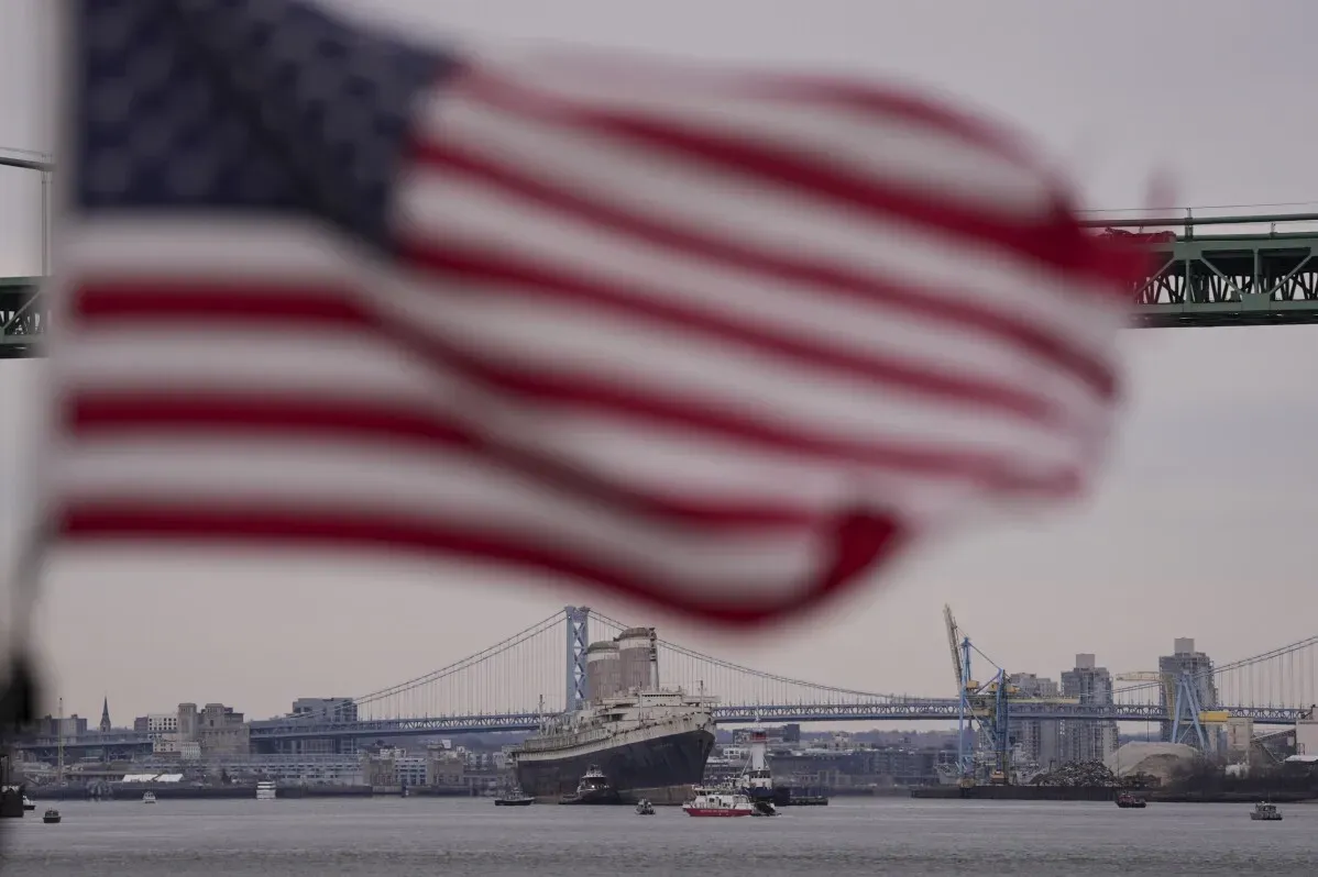 سفينة SS United States التاريخية تبحر في نهر ديلاوير، مع علم أمريكي يرفرف في المقدمة، استعدادًا لتحويلها إلى شعاب مرجانية اصطناعية.