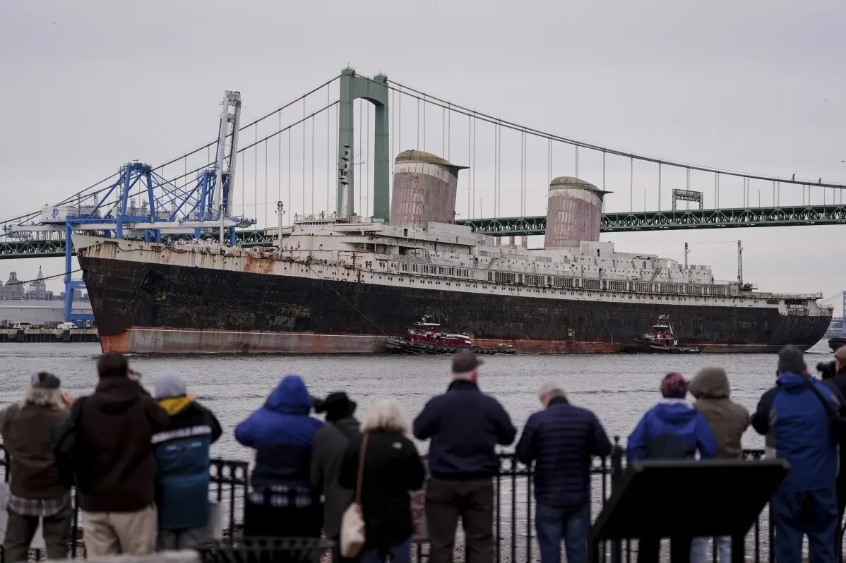 سفينة SS United States التاريخية تبحر من نهر ديلاوير، مع جسر خلفها ومشاهدين يتابعون الرحلة نحو تحويلها إلى شعاب مرجانية اصطناعية.