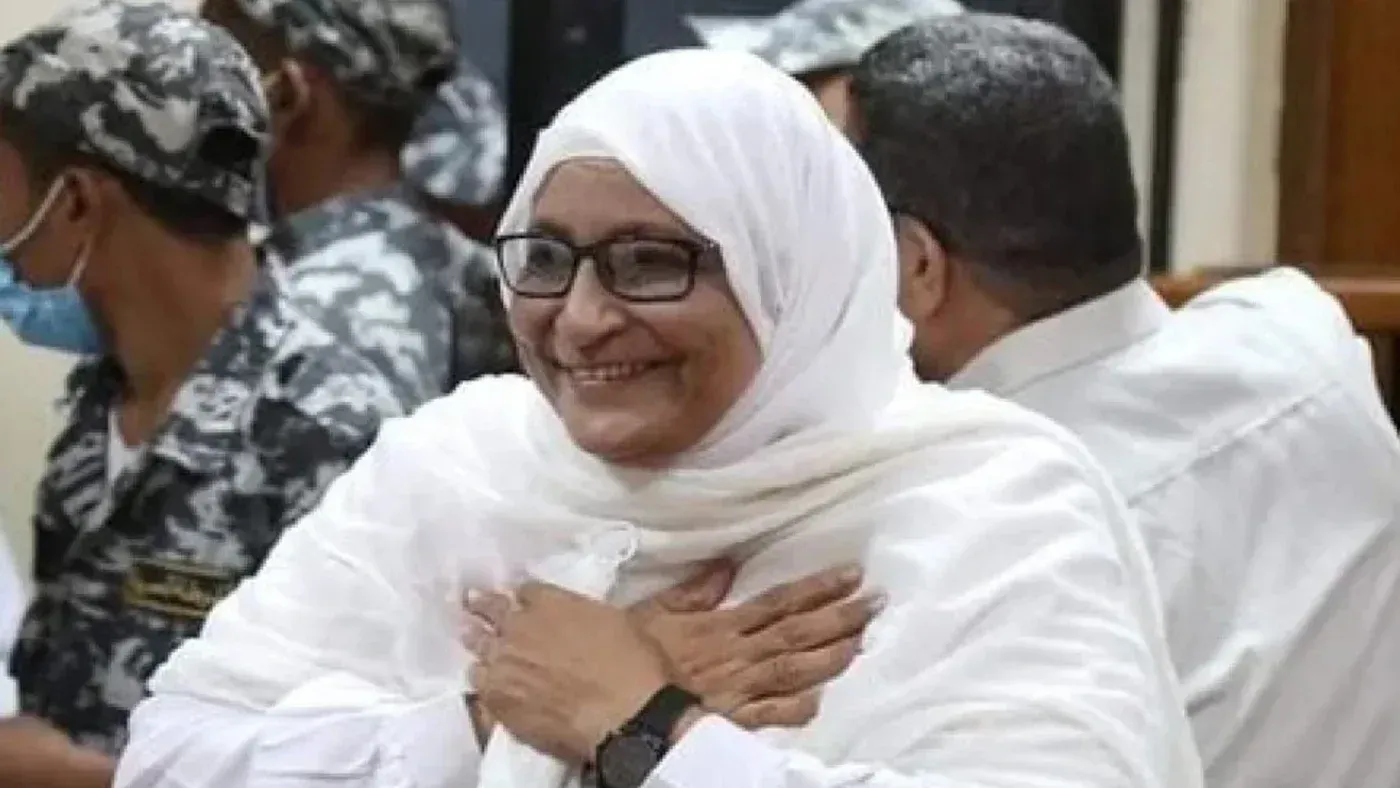 امرأة مسنّة ترتدي الحجاب الأبيض، تبتسم في قاعة المحكمة، محاطة بمجموعة من رجال الأمن بزيّ رسمي.