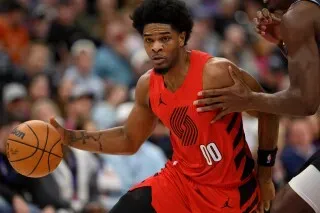 جونيور هوليداي يحمل كرة السلة أثناء المباراة، مرتديًا زي بورتلاند تريل بليزرز، مع وجود لاعب آخر بالقرب منه في خلفية الملعب.