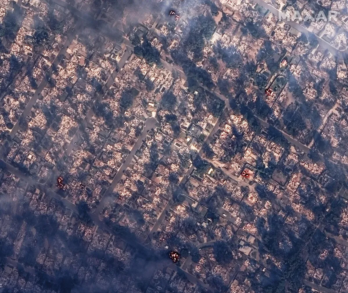 تظهر الصورة مشهدًا جويًا لمناطق مدمرة بسبب حرائق لوس أنجلوس، حيث تحولت المنازل إلى أنقاض وسط دخان كثيف.