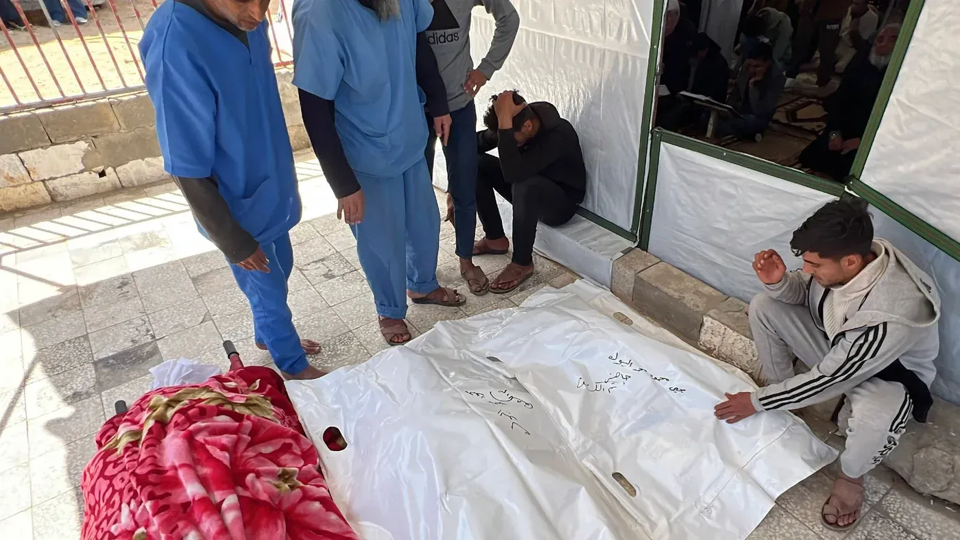 مجموعة من الرجال يرتدون ملابس طبية زرقاء يتجمعون حول جثة مغطاة بغطاء أبيض، في سياق مأساوي يعكس آثار الإبادة الجماعية في غزة.