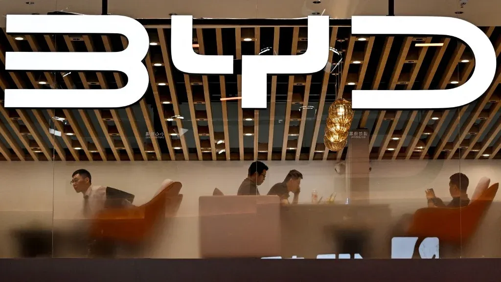 شعار شركة BYD يظهر في واجهة مكتب حديث، مع موظفين يعملون في الداخل، مما يعكس التوجه نحو إنتاج السيارات الكهربائية.