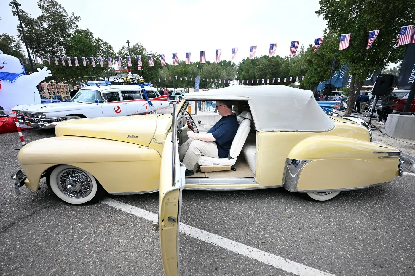 سيارة كلاسيكية صفراء مكشوفة متوقفة في حدث عام، مع شخص يجلس داخلها، وخلفها سيارات أخرى وزينة بأعلام أمريكية.