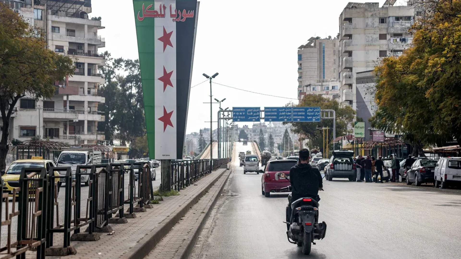 افتتاحية لمدينة سورية تظهر لافتة كبيرة تحمل نجوم حمراء على خلفية العلم الوطني، مع مشهد حضري يتضمن سيارات وأشخاص في الشارع.