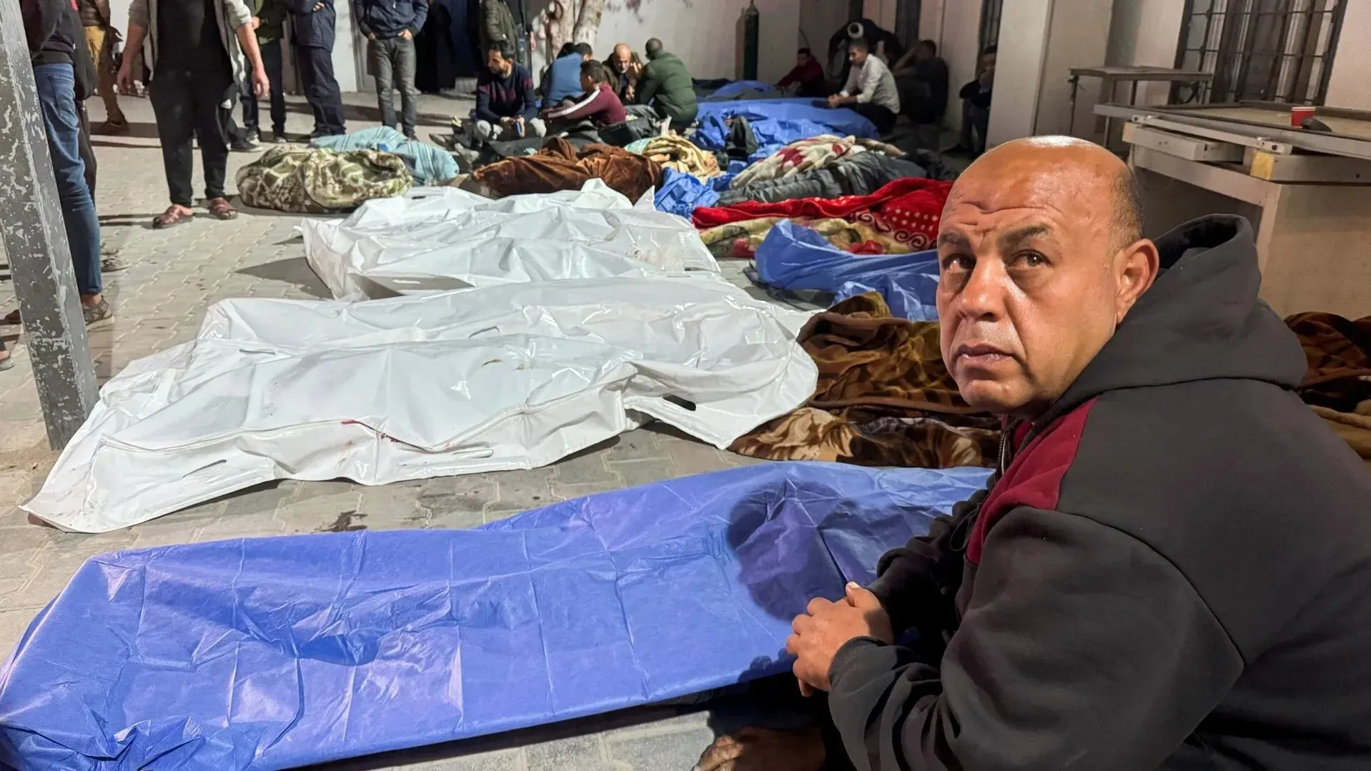 رجل يجلس في مكان مخصص للضحايا، محاط بأغطية وأشخاص آخرين، بعد الهجمات الإسرائيلية الأخيرة على غزة التي أسفرت عن مئات القتلى.