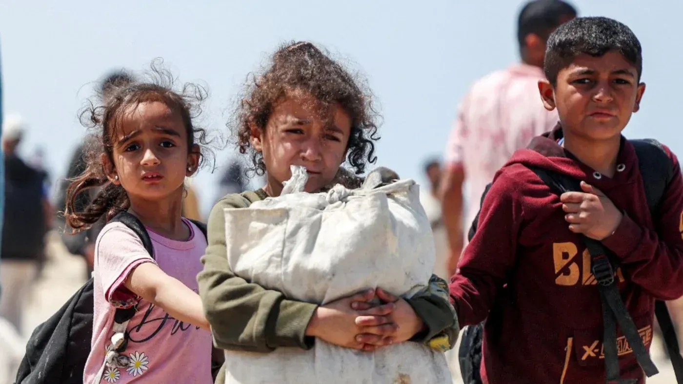 أطفال فلسطينيون نازحون يحملون أمتعتهم، يعبرون مناطق مدمرة في غزة، يعكسون معاناة النزوح وفقدان الهوية في ظل الحرب.