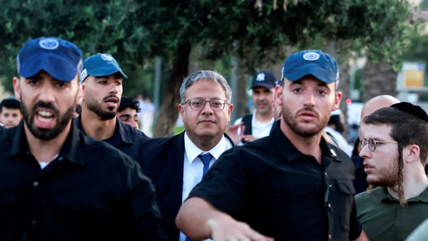 رجل يرتدي بدلة مع حراس أمن خلال حدث عام، محاط بأشخاص آخرين، في سياق توترات سياسية في القدس.