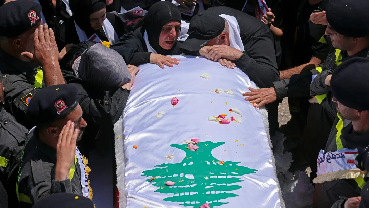 تجمع حشود من الناس حول نعش مغطى بعلم لبنان، حيث يعبرون عن حزنهم بفقدان أحد الشهداء، مع تواجد عناصر من الأمن في الخلفية.