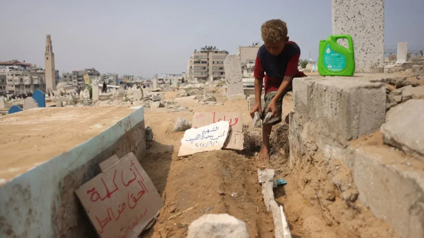 طفل صغير يعمل في مقبرة في غزة، يحفر في الأرض بالقرب من شواهد القبور، مع وجود لافتات مكتوبة باللغة العربية.