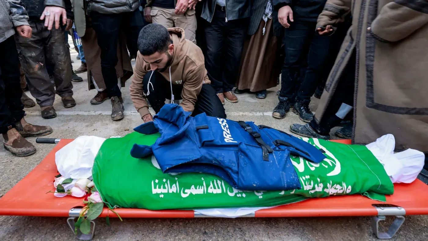 جثمان صحفية فلسطينية مغطاة بعلم أخضر، مع سترة صحفية زرقاء، محاطة بأشخاص في مشهد يعبّر عن الحزن بعد الهجمات الإسرائيلية.