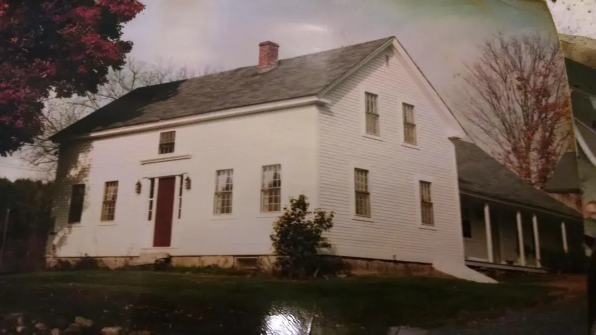 صورة لمنزل قديم ذو جدران بيضاء وسقف رمادي، يحيط به حديقة صغيرة وأشجار، تجسد تاريخ العائلة.