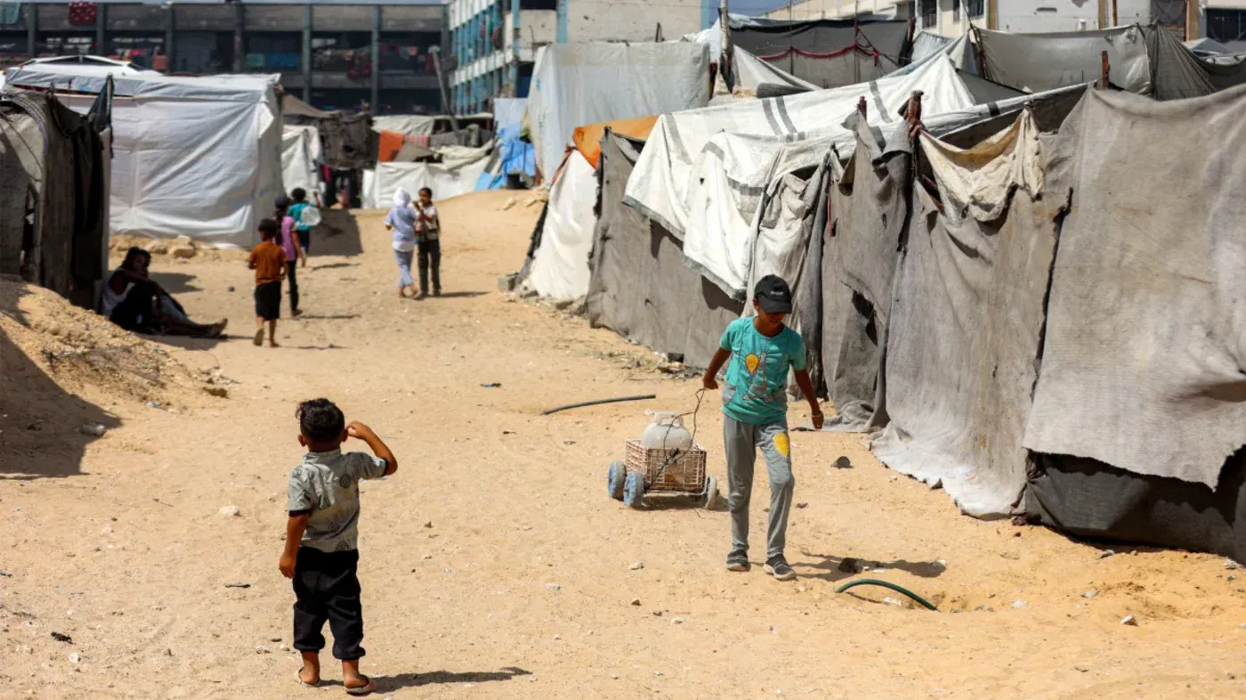 أطفال يلعبون في مخيم للاجئين الفلسطينيين، يظهرون بين الخيام البيضاء والرمادية في بيئة قاحلة، تعكس معاناة اللاجئين.