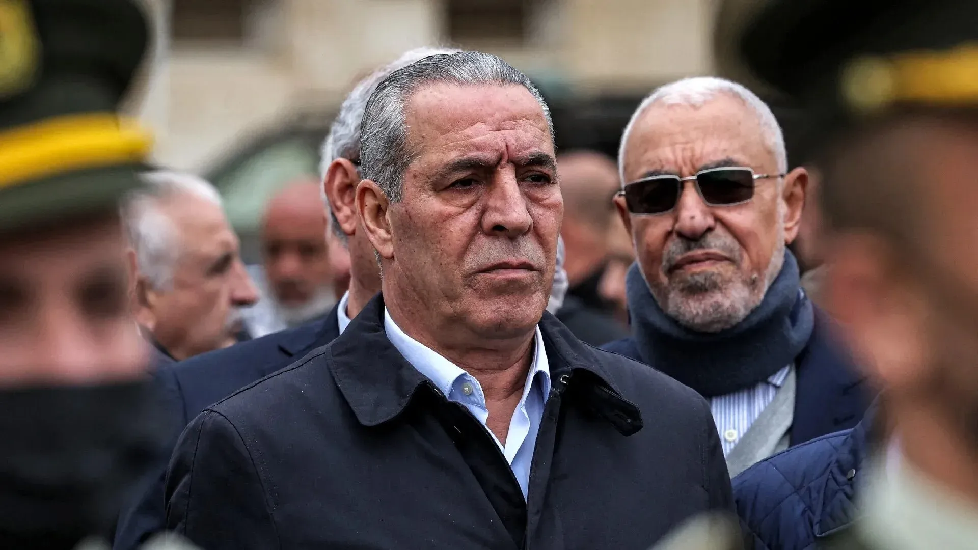 حسين الشيخ، نائب الرئيس الفلسطيني الجديد، يظهر في حدث رسمي، مع خلفية من رجال أمن ومساعدين، مما يعكس دوره السياسي البارز.