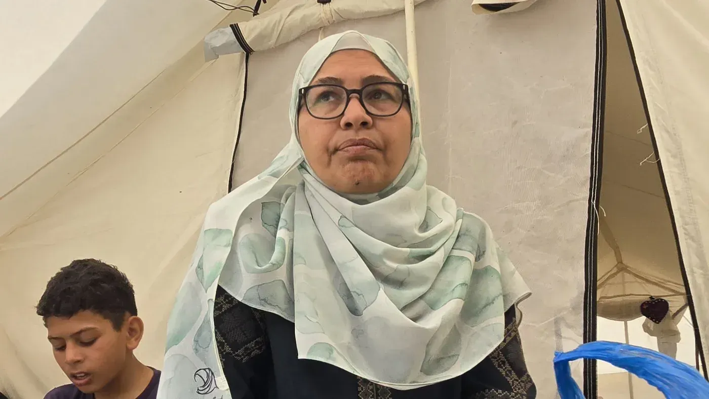 امرأة ترتدي حجاباً وتبدو قلقة، تجلس داخل خيمة في مخيم للنازحين، بينما يظهر طفل صغير بجانبها، يعكس معاناة العائلات في غزة.