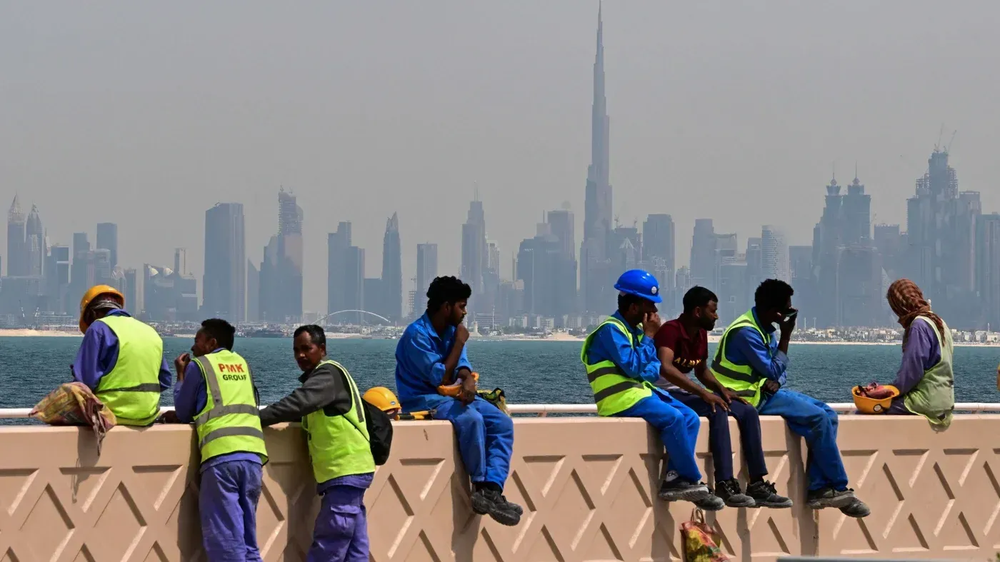 مجموعة من العمال المهاجرين يرتدون زي العمل، يجلسون على حافة جسر في دبي، مع skyline المدينة في الخلفية، تعبيرات وجوههم تعكس التحديات التي يواجهونها.