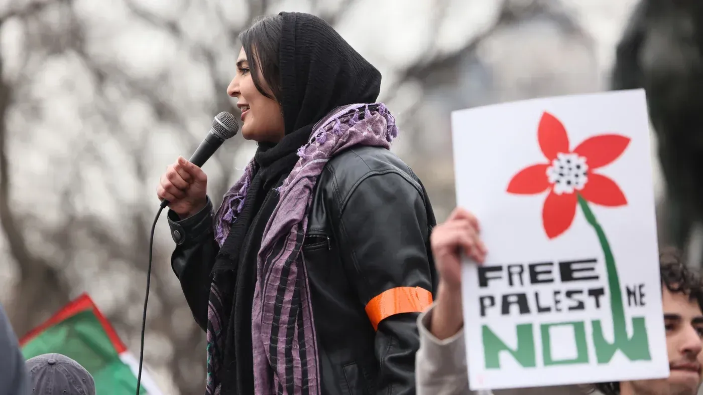 ناشطة فلسطينية-أمريكية تتحدث في مظاهرة، تحمل لافتة تطالب بتحرير فلسطين. تظهر خلفها أعلام فلسطينية وأجواء احتجاجية.