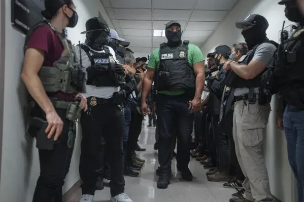 رجال شرطة يرتدون سترات واقية وأقنعة، يقفون في ممر ضيق، في سياق عمليات إنفاذ قوانين الهجرة والجمارك الأمريكية.