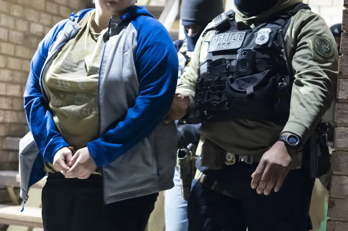 اعتقال شخص أثناء عملية تنفيذ قوانين الهجرة، حيث يقوم ضابط يرتدي سترة مضادة للرصاص بإمساك ذراعه في بيئة حضرية.