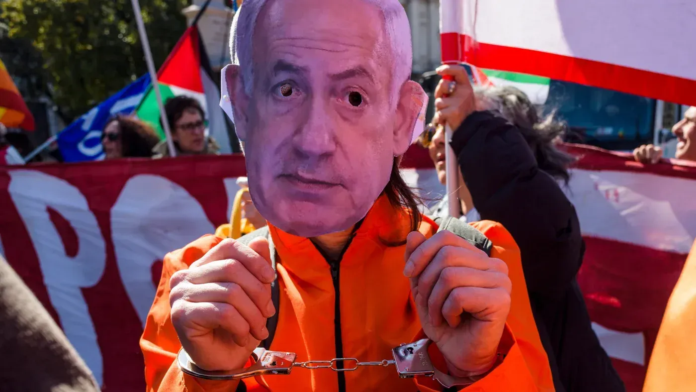 شخص يرتدي زيًا برتقاليًا ويمسك بأصفاد، يحمل قناعًا يمثل رئيس الوزراء الإسرائيلي بنيامين نتنياهو، وسط مظاهرة.