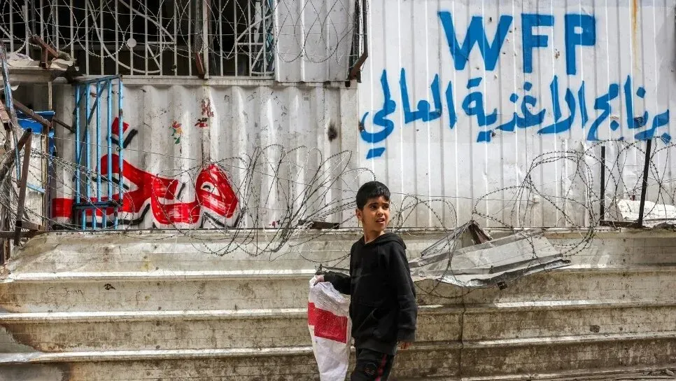 طفل فلسطيني يقف بجوار حواجز شائكة أمام مركز برنامج الأغذية العالمي، مع وجود كتابات تشير إلى الحاجة الماسة للمساعدات الغذائية في غزة.