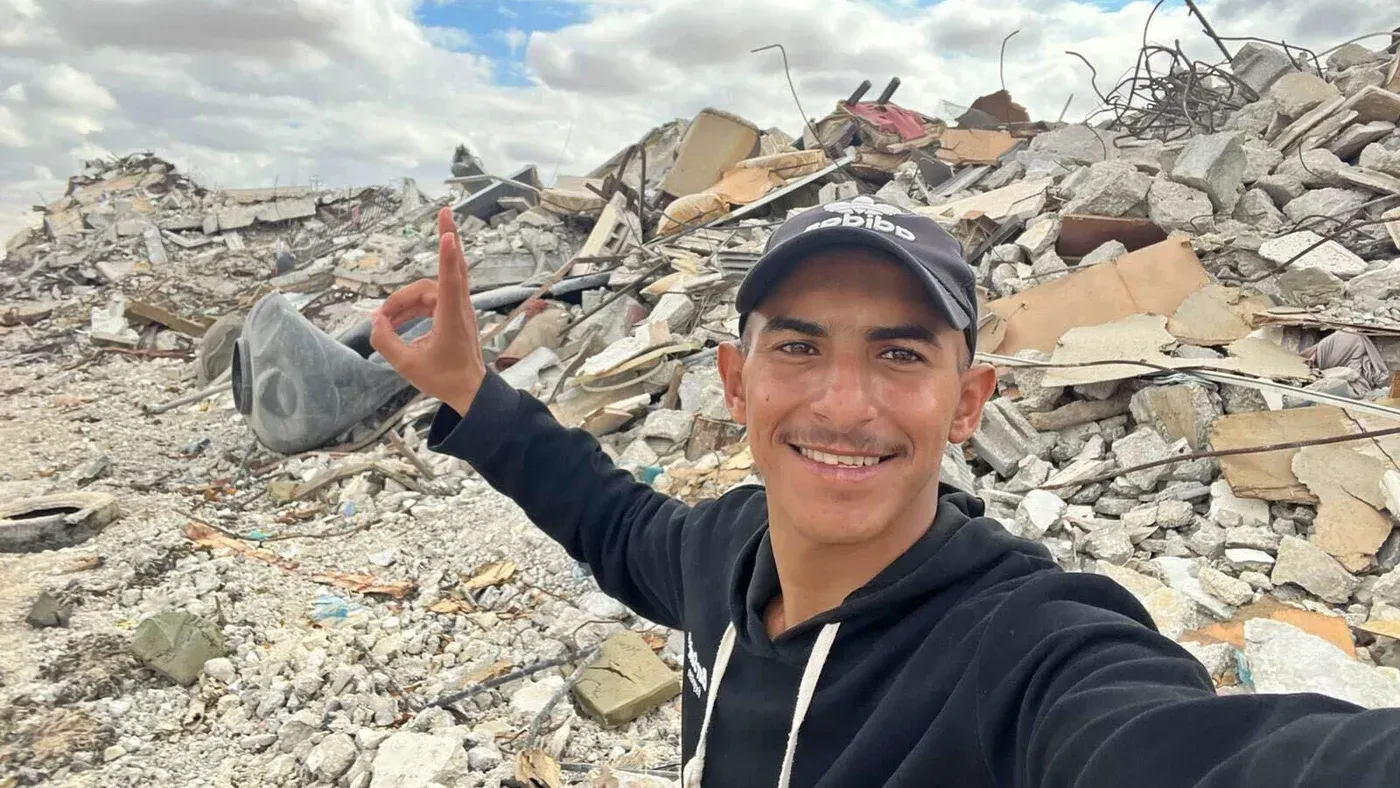 شاب مبتسم يقف أمام أنقاض مبنى مدمر، مع رفع إصبعه في إشارة النصر، تعبيرًا عن الأمل وسط الدمار في غزة.
