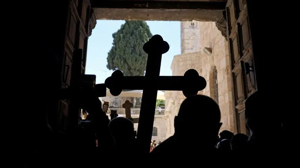 صورة لمجموعة من المسيحيين الفلسطينيين يحملون الصليب عند مدخل كنيسة القيامة في القدس، مع خلفية تظهر الشجرة والأبنية.