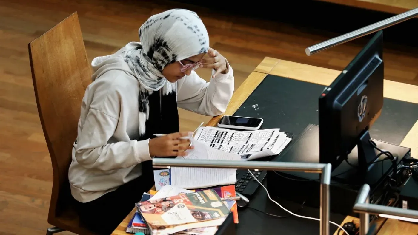 شابة ترتدي حجابًا وتدرس في مكتبة، محاطة بمجموعة من الكتب والأوراق، وتظهر في خلفية حاسوب وأجهزة دراسية، تعكس تحديات التعليم في مصر.