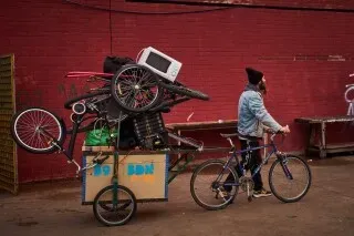 رجل يدفع عربة محملة بأغراض متنوعة تشمل دراجات وأجهزة كهربائية، في خلفية حائط أحمر، يعكس جهود إعادة التدوير في المجتمع.