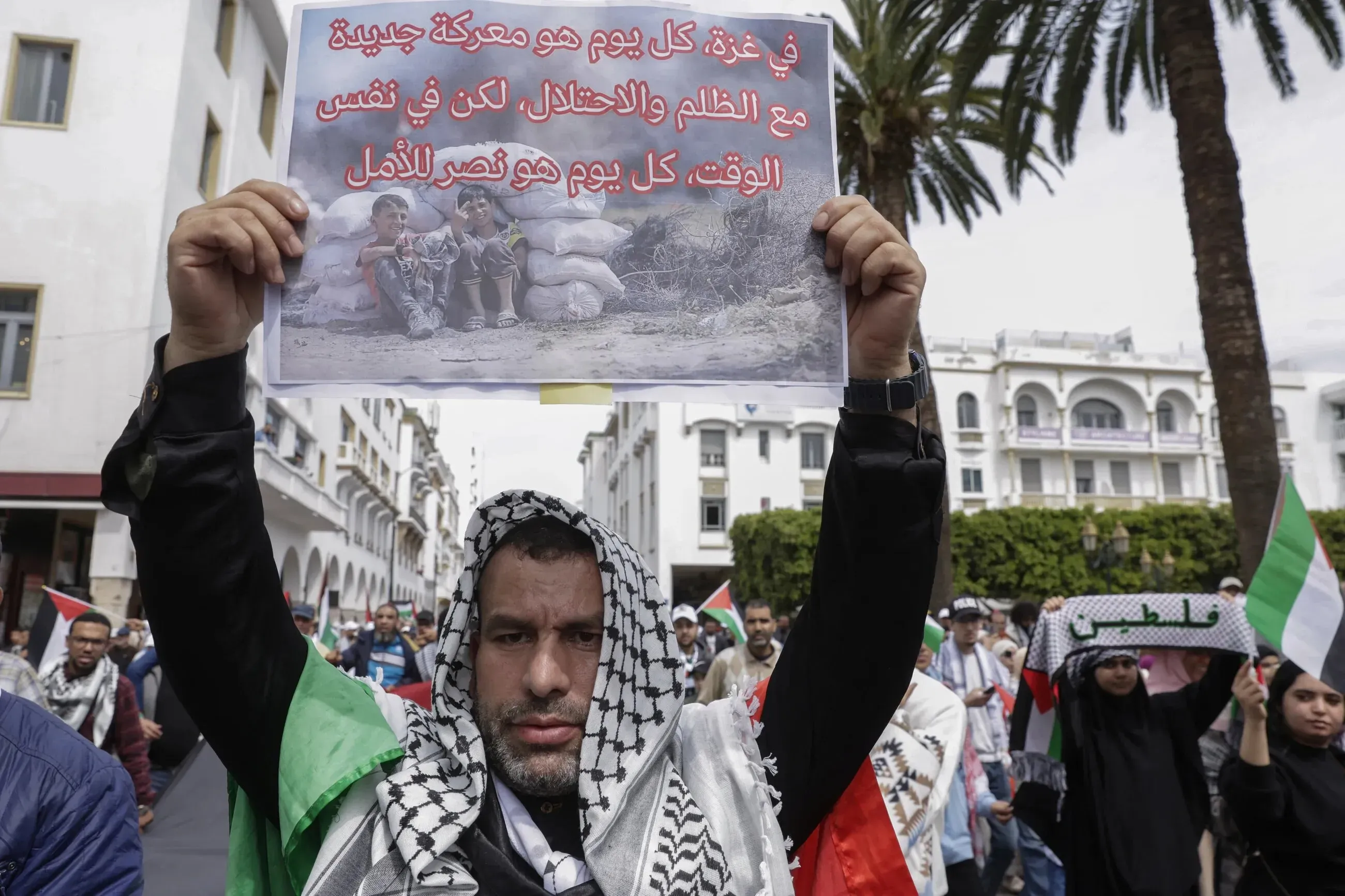 رجل يحمل لافتة في مظاهرة، تعبر عن معاناة غزة وتطالب بالعدالة، مع خلفية لأشخاص آخرين يحملون الأعلام الفلسطينية.