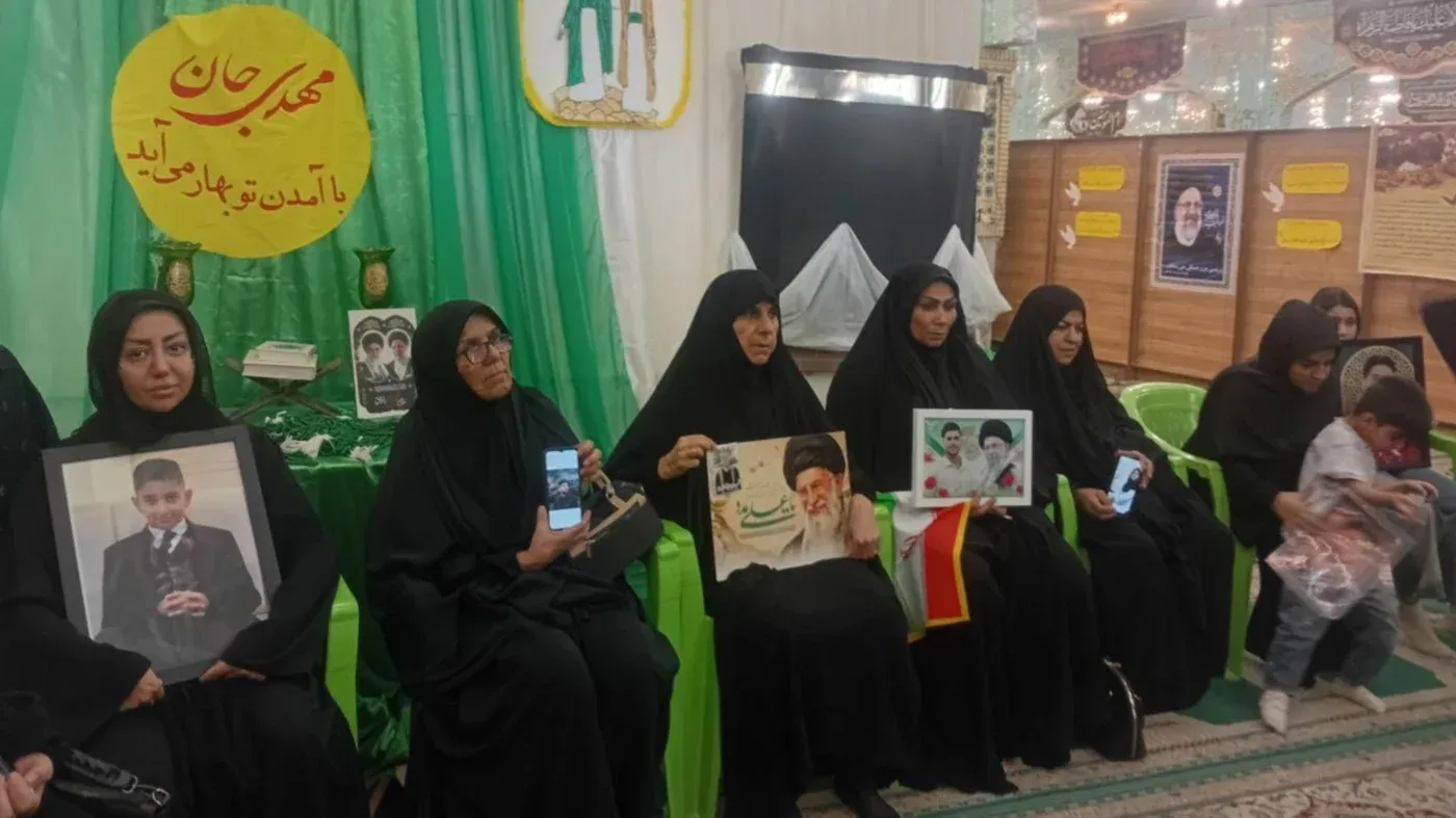 نساء يرتدين الحجاب الأسود يجلسن في قاعة، يحملن صورًا لأحبائهن، مع خلفية مزينة بالأخضر، تعبيرًا عن الحزن بعد الهجوم على لامرد.