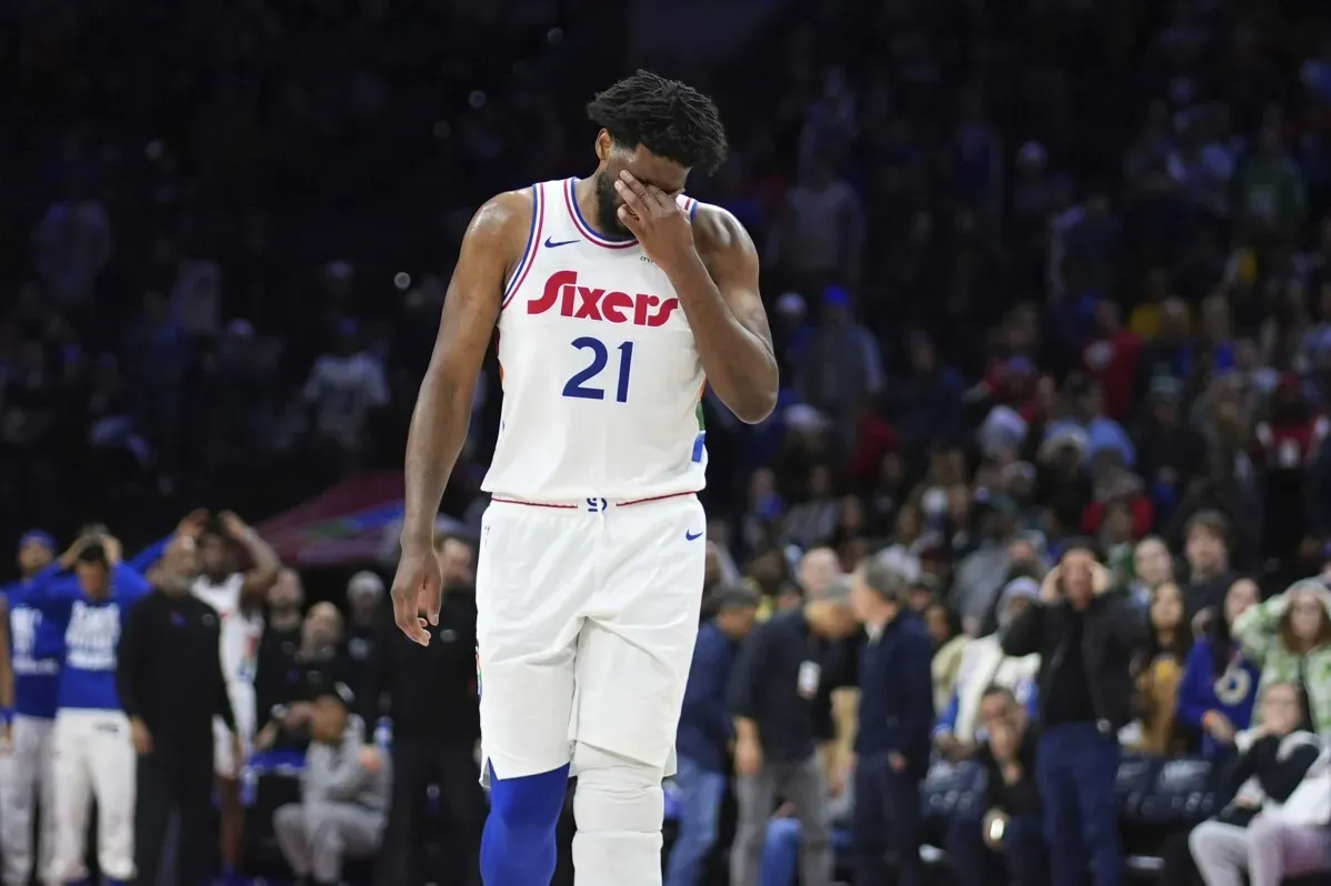 جويل إمبيد، لاعب فريق 76ers، يبدو محبطًا بعد خسارة المباراة، مع وجود مشجعين في الخلفية. تعكس الصورة معاناته بسبب الإصابات المتكررة.