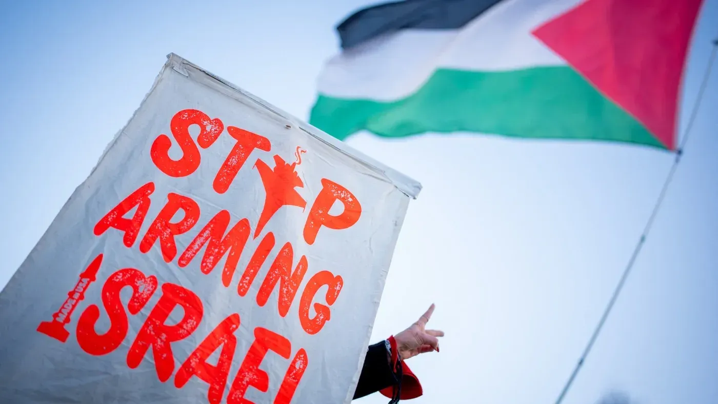 لافتة تحمل عبارة "أوقفوا تسليح إسرائيل" مع علم فلسطين في الخلفية، تعكس دعوات لإنهاء الدعم العسكري لإسرائيل.