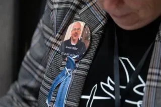 شخص يرتدي سترة مزينة بشارة تحمل صورة شخص، خلال جلسة استماع حول حادث تحطم طائرة، تعبير عن الحزن والذكرى للضحايا.