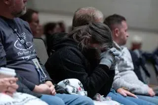 امرأة ترتدي سترة سوداء وتغطي وجهها بيديها أثناء جلسة استماع حول حادث تحطم الطائرات، بينما يجلس آخرون في الخلف.