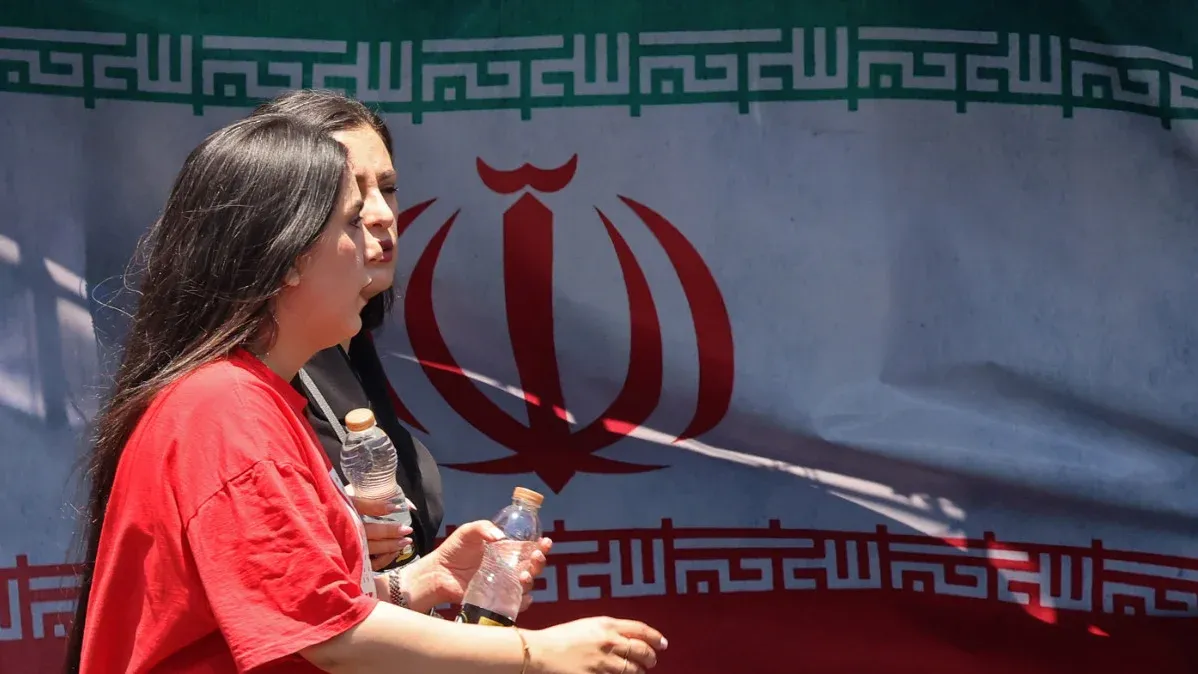 امرأتان تسيران أمام علم إيران، يحملن زجاجات مياه، تعبيراً عن الأمل في تحقيق اتفاق سلام بين إيران والولايات المتحدة.