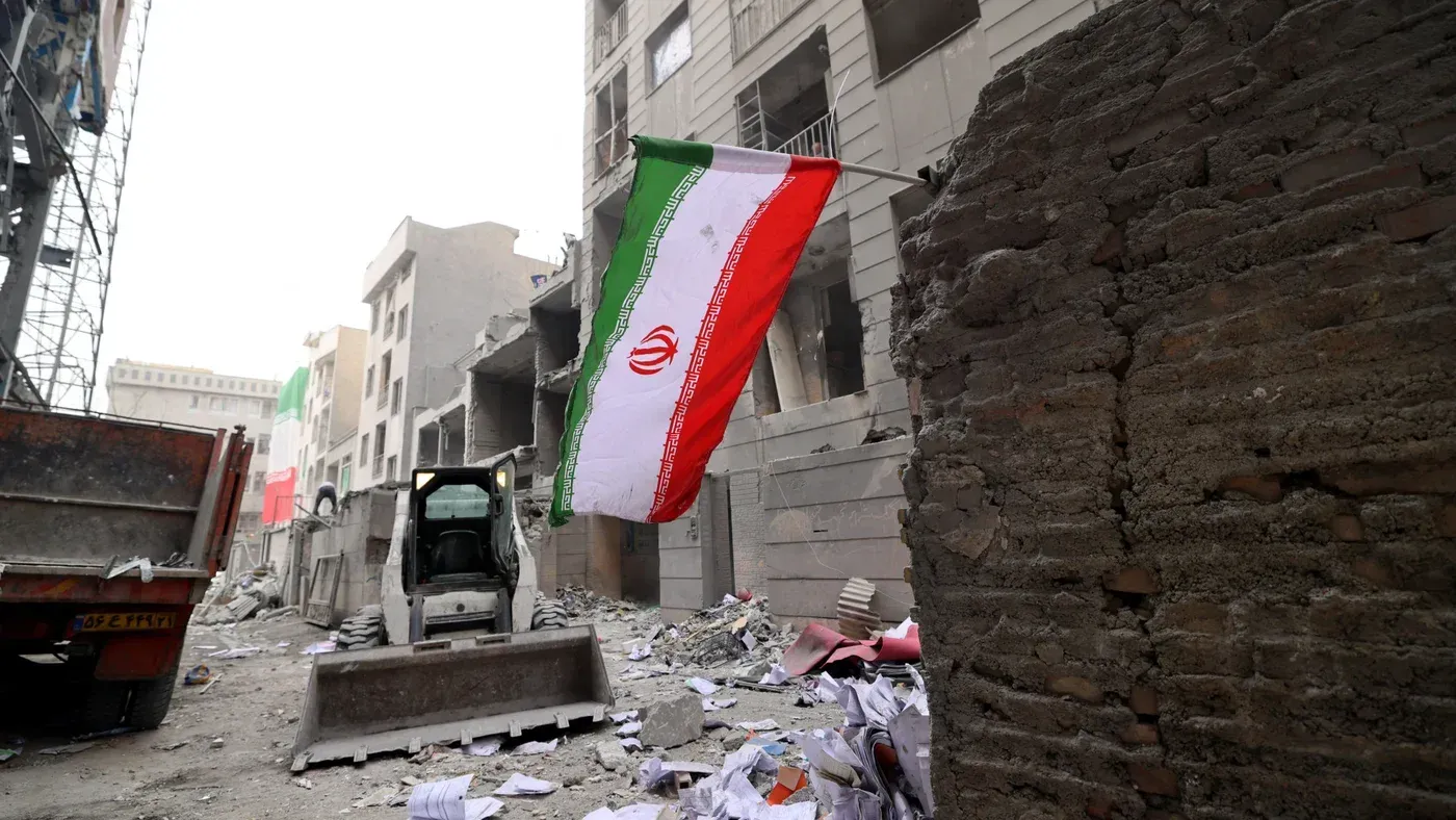 علم إيران يرفرف في موقع دمار، مع وجود حطام المباني وآثار الدمار حوله، مما يعكس تأثير الغزو المستمر على البلاد.