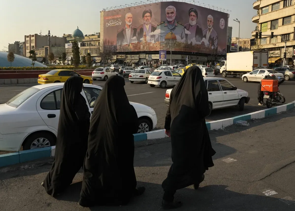 نساء يرتدين الحجاب الأسود يسيرن في شوارع طهران، خلفهن لافتة كبيرة تحمل صور شخصيات سياسية، وسط حركة المرور.