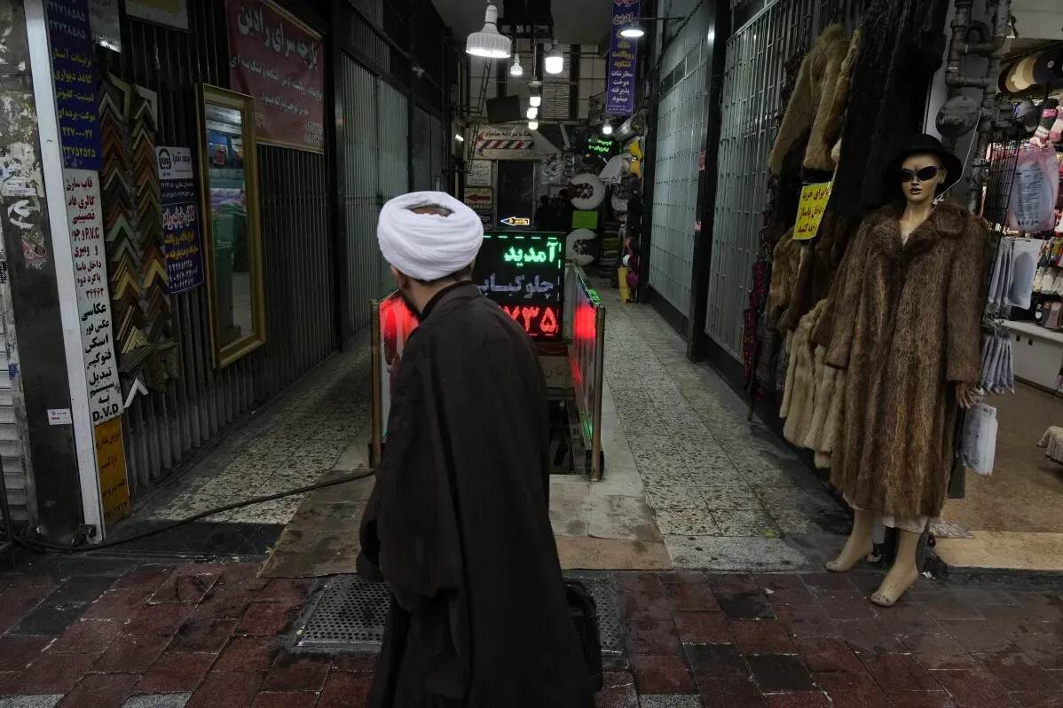رجل يرتدي عمامة وعباءة يسير في سوق إيراني، بينما تعرض دمى ملابس على اليمين، مما يعكس التوتر بين القيم التقليدية والحداثة.