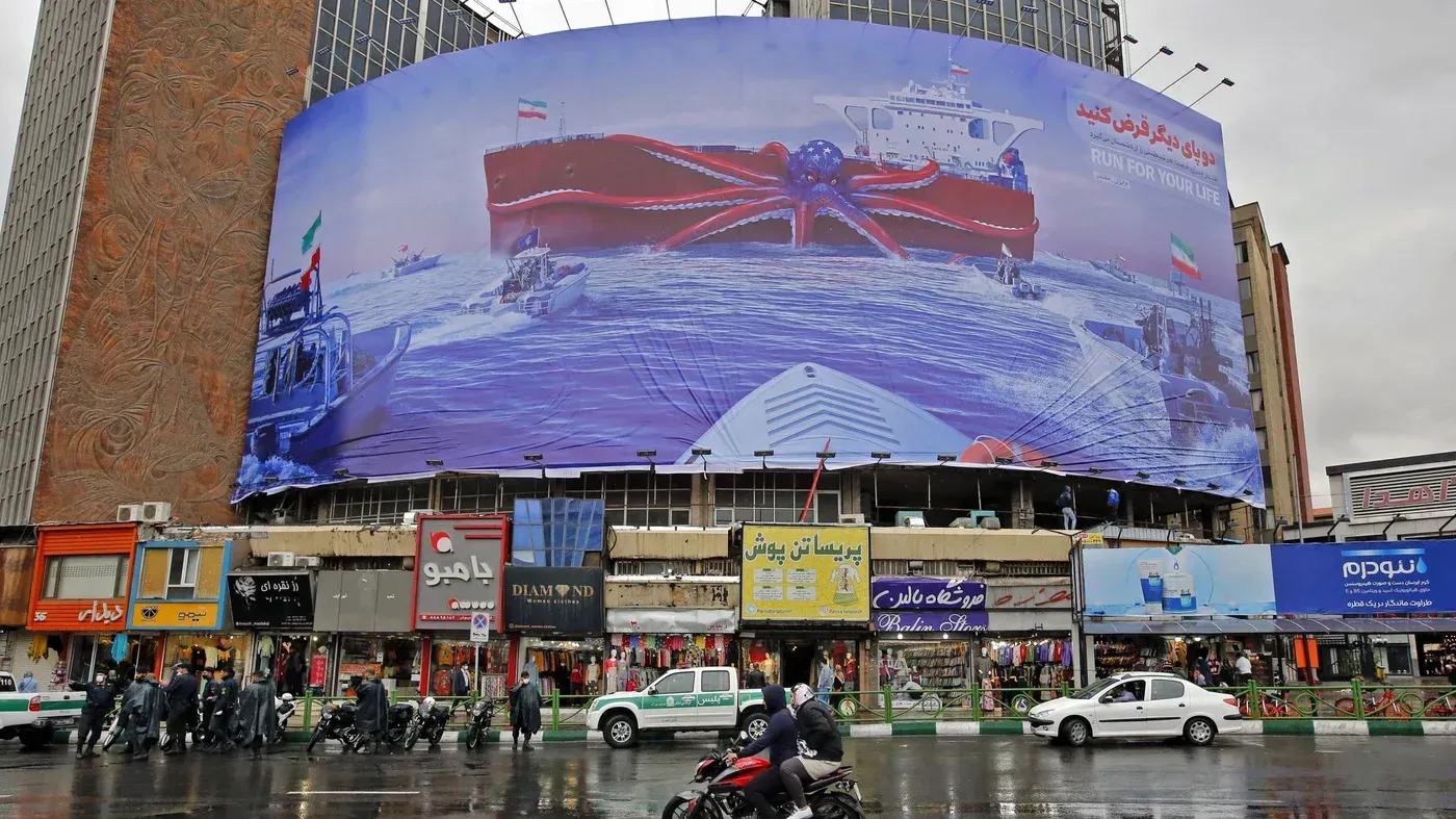 صورة لافتة إعلانية ضخمة تظهر ناقلة نفط حمراء في البحر، مع زوارق حراسة بالقرب منها، تعكس التوترات البحرية في الخليج.