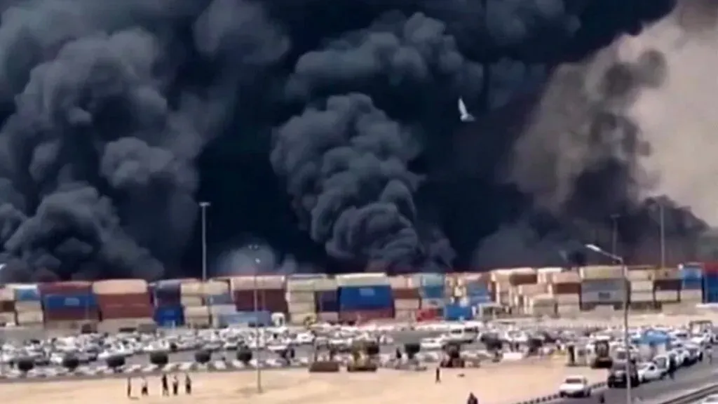تصاعد الدخان الكثيف من ساحة سينا للحاويات في بندر عباس بعد انفجار كبير، مع وجود مركبات ومباني متضررة في المنطقة.