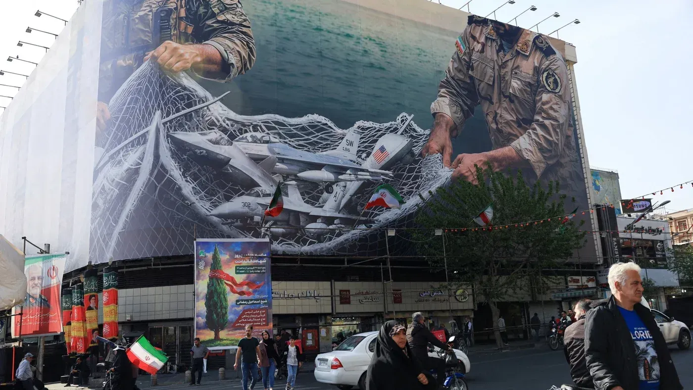 لوحة إعلانات ضخمة في طهران تُظهر جنديًا إيرانيًا يمسك بشبكة تحتوي على طائرات مسيرة، مع العلم الإيراني في الخلفية، تعكس التوترات الإقليمية.