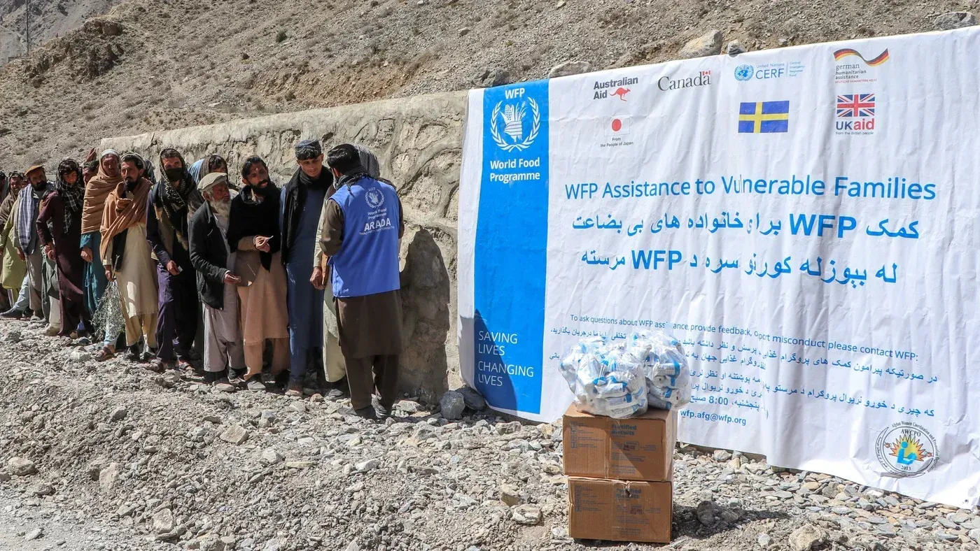 طابور من الأشخاص ينتظرون المساعدات الغذائية من برنامج الأغذية العالمي، مع لافتة توضح الدعم المقدم للعائلات الضعيفة.