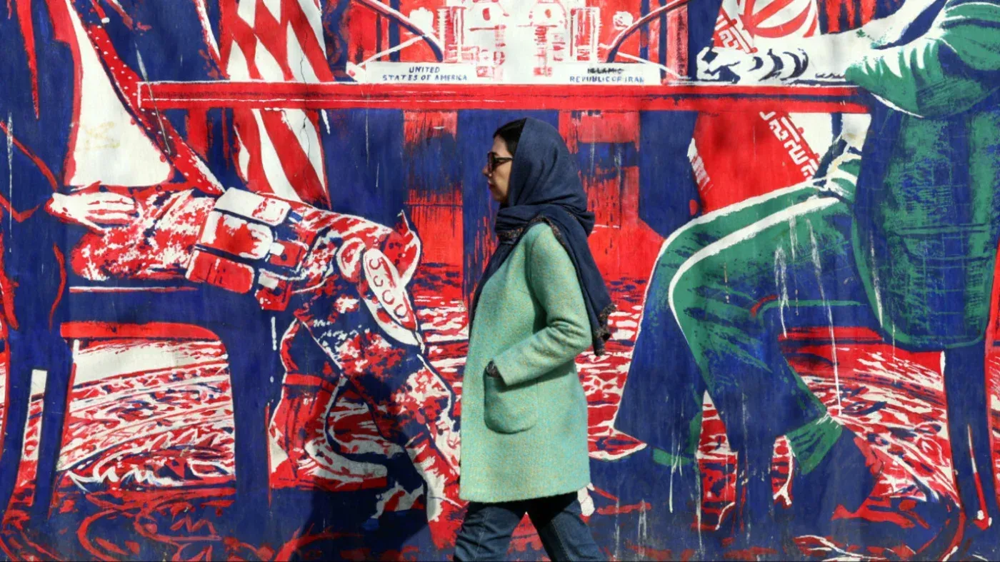 امرأة ترتدي معطفًا أخضر تسير بجانب جدارية ملونة تمثل العلاقات الإيرانية الأمريكية، تعكس التوترات السياسية الحالية.