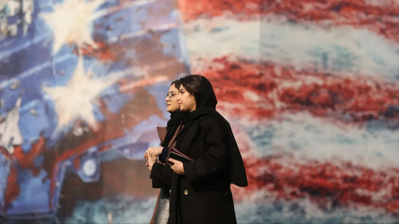 امرأتان ترتديان الحجاب تسيران معًا أمام جدارية ملونة تتضمن ألوان العلم الأمريكي، تعكس الأجواء الاجتماعية والسياسية في إيران.