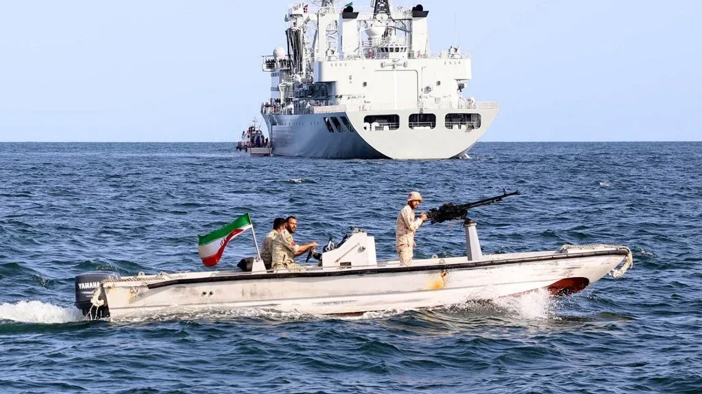 زورق حرس ثوري إيراني مسلح يبحر في الخليج الفارسي مع سفينة حربية كبيرة في الخلفية، يعكس استراتيجية إيران البحرية في مضيق هرمز.