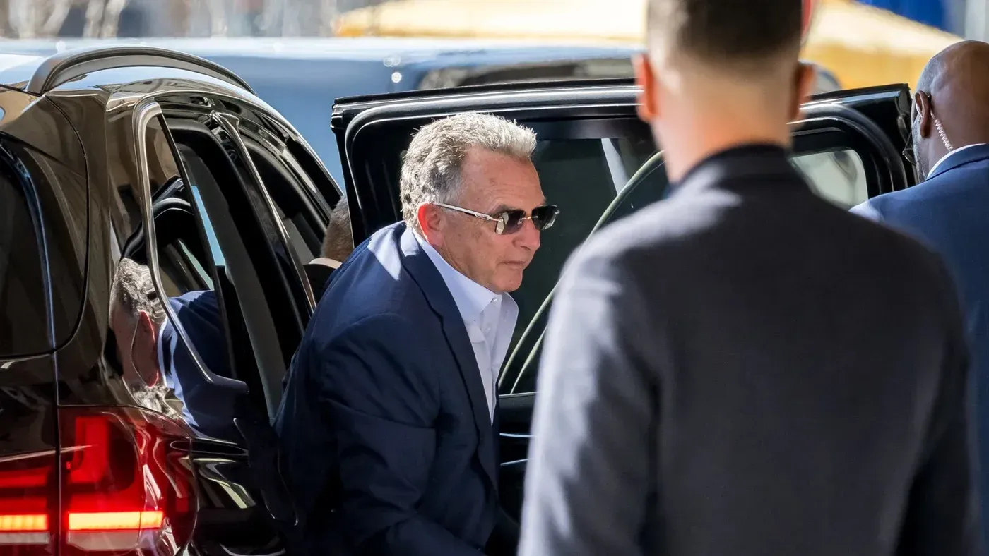 رجل يرتدي نظارات شمسية يخرج من سيارة سوداء، محاطًا بأشخاص آخرين، مما يعكس أجواء من الحذر والاهتمام الأمني.