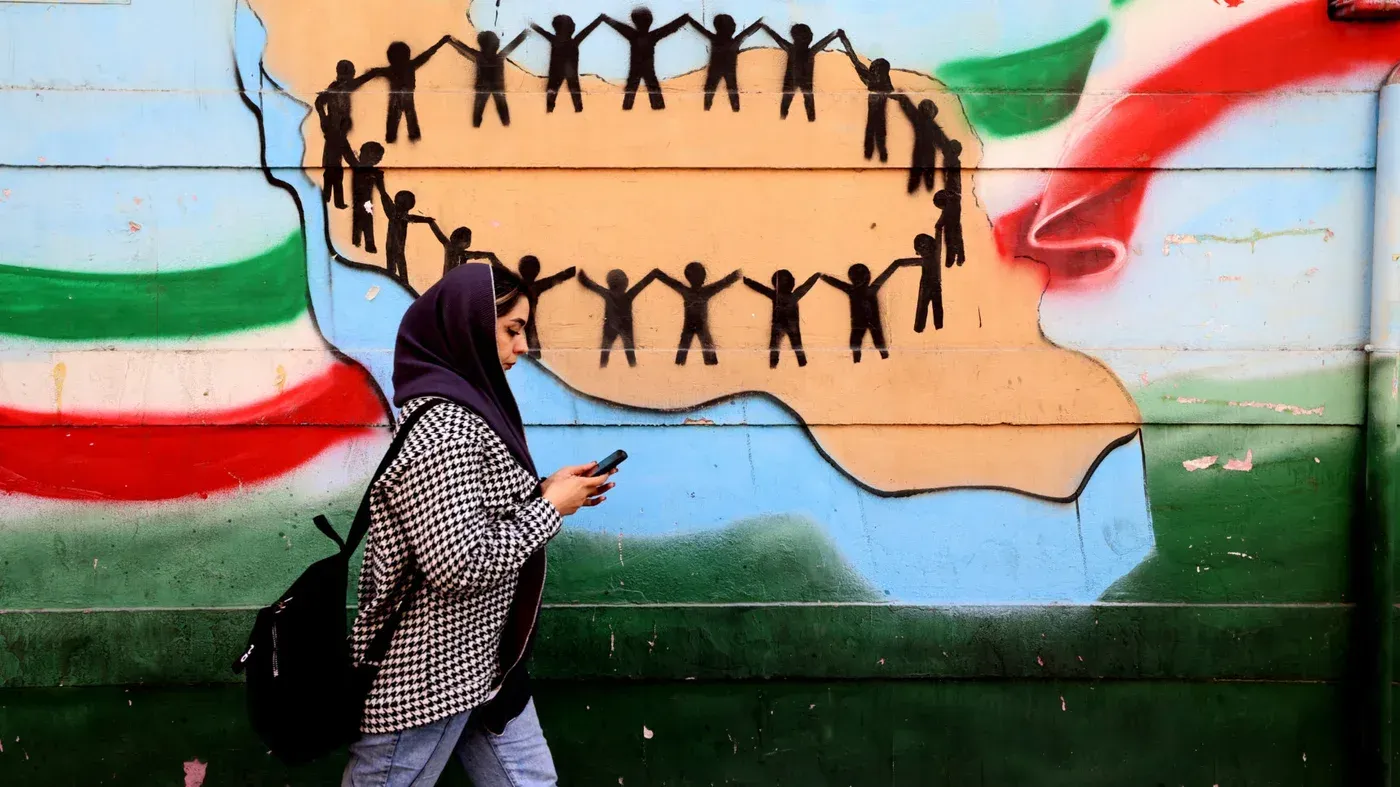 امرأة ترتدي حجابًا تسير بجوار جدارية ملونة تمثل التضامن، مع أشخاص يتشابكون بأيديهم على شكل خريطة إيران.