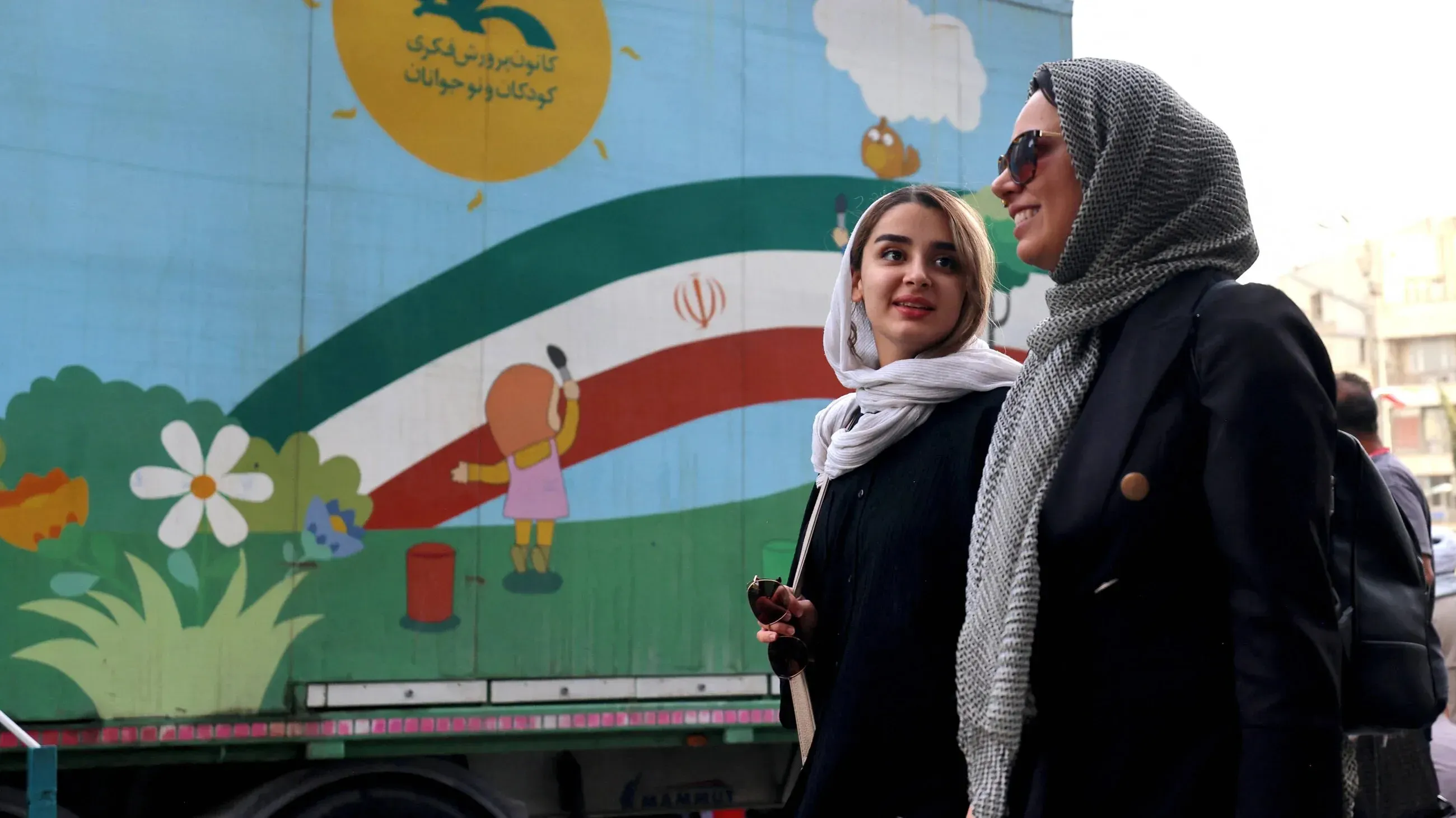 امرأتان ترتديان الحجاب يتحدثان معًا في الشارع، بينما يظهر خلفهما جدار مزين برسوم ملونة تعكس الثقافة الإيرانية.