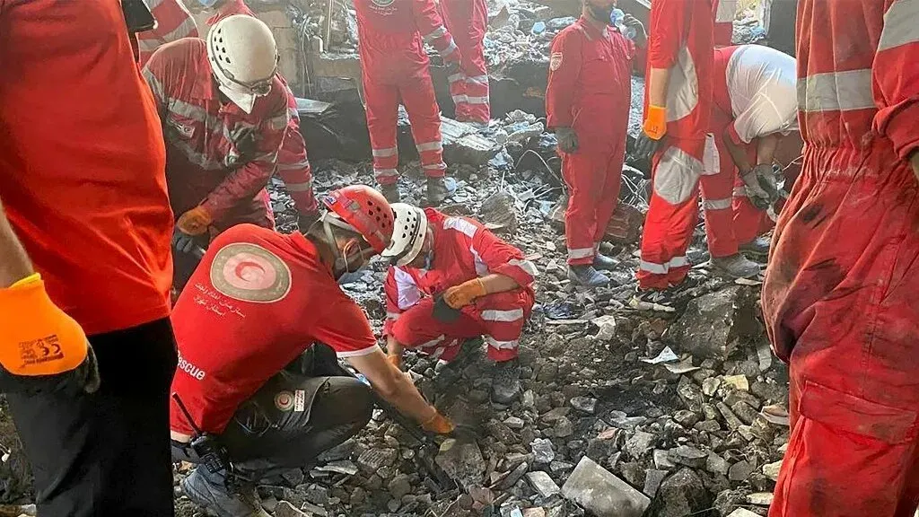 عمال إنقاذ يرتدون زيًا برتقاليًا وأحمر، يبحثون بين الأنقاض بعد قصف في طهران، وسط فوضى وقلق متزايد بين المدنيين.